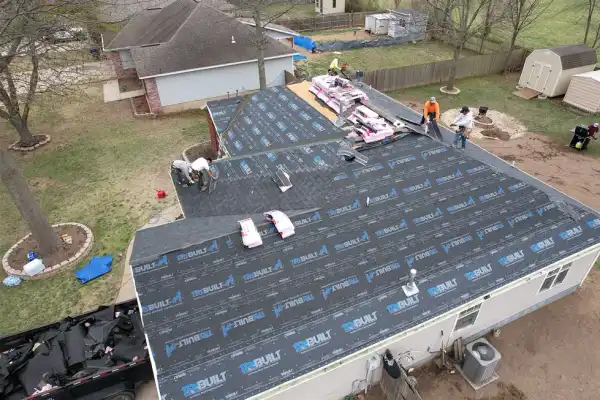 Drone view of a crew laying new shingles over black underlayment on a long ranch roof, bundles staged along the ridge