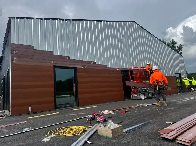 Crew in hi-vis installing wood-look metal wall panels on a new steel building, with a scissor lift and materials on the slab