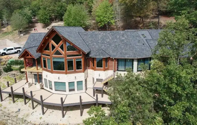 Drone view of a log and stone lake home on a wooded bluff with a new dark slate-look shingle roof and a Roofing & Gutter Experts truck on the drive